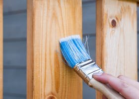 A hand painting a wooden fence plank with a blue paintbrush outdoors. Perfect for DIY and renovation themes.