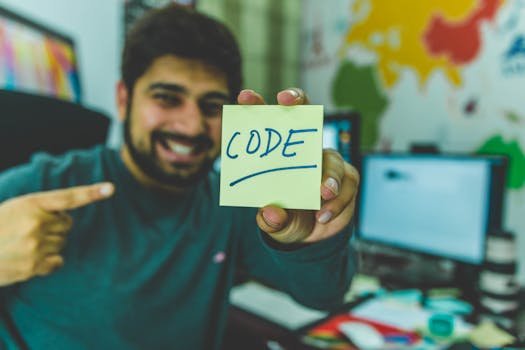 Game development A smiling Game Development programmer pointing to a sticky note with 'code' written on it in an office setting.