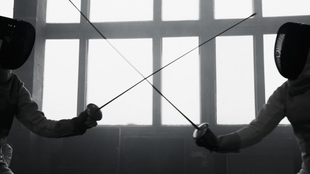 Two fencers engage in a dramatic indoor match, crossing swords in a black and white setting.